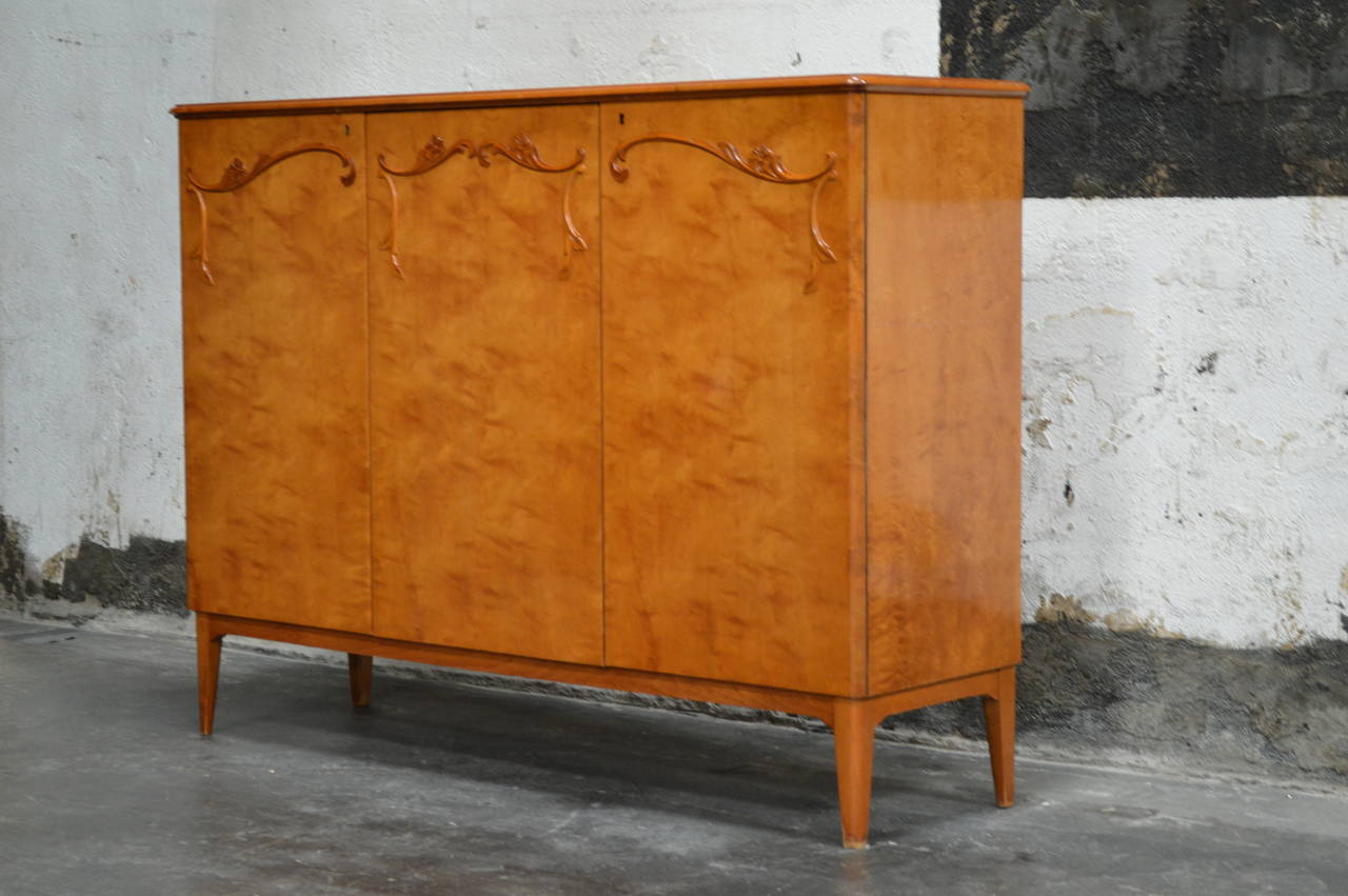 Art moderne cabinet crafted of native golden flame birch with carved floral details. This gorgeous wood has mellowed to a rich amber color. This storage piece opens to reveal a golden elm interior with deep shelves and three drawers. Key included.