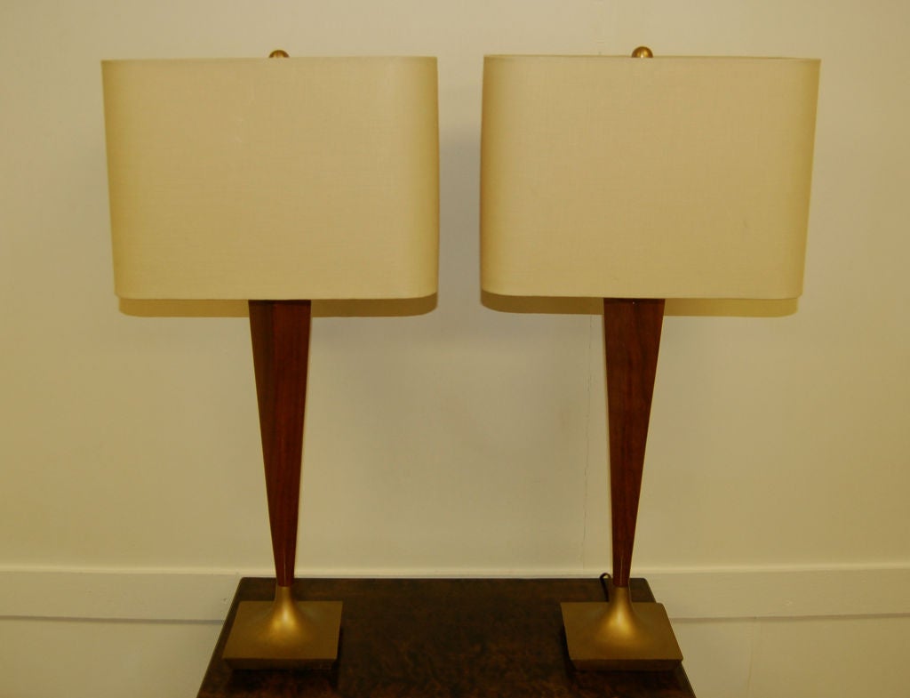 Stylized pair of teak and brass table lamps.  Sleek tapered walnut lamps on square cast brass bases.  Newly rewired to US standards.  New curved rectangular almond linen shades.  Solid brass ball finials.