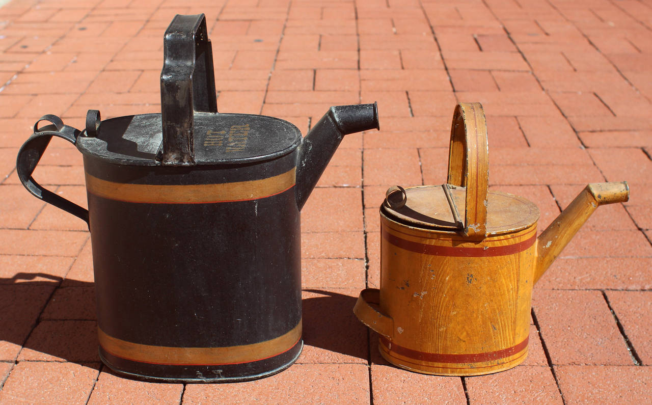 Two watering cans - each painted galvanized steel, one black with gold and red line decoration and the other in a faux wood with red line decoration. Both read 