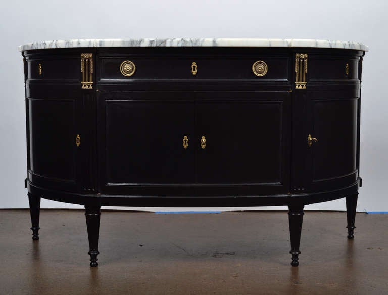 Antique French Louis XVI style demilune buffet with a  Carrara marble top. Strongly constructed of mahogany, ebonized with a glossy French polish finish. Four locking doors and a single dovetailed drawer with fine lost wax bronze hardware and