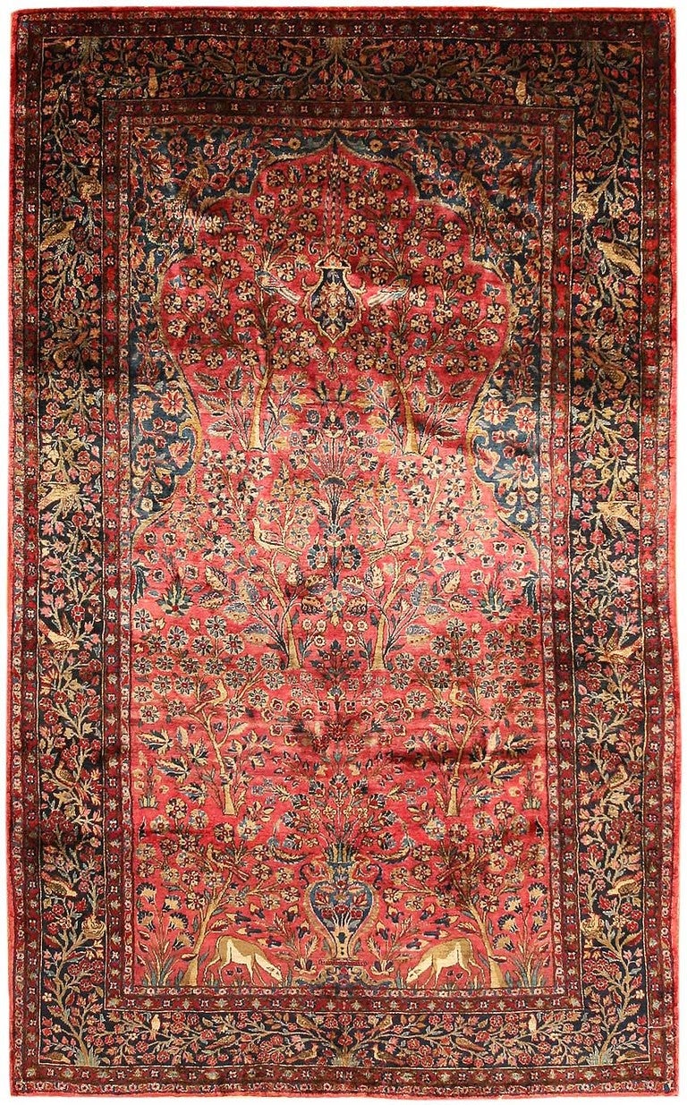 Featuring a central vase flanked by grazing deer, this pastoral Kashan carpet is woven with a rich burgundy background decorated with millefleur-style clusters of flowering trees and dense foliage that provides habitat for a multitude of auspicious