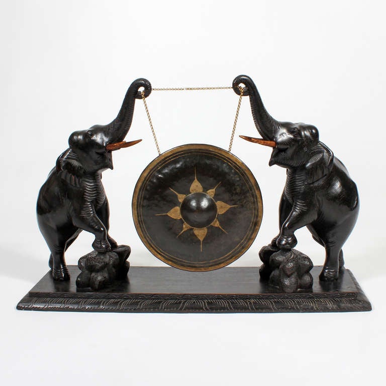 This is a very large and impressive carved and ebonized teak elephant gong. Two elephants, trunks raised, standing on rocks, flank a large decorated brass gong, all mounted on a carved platform base. Complete with the original gong, this piece