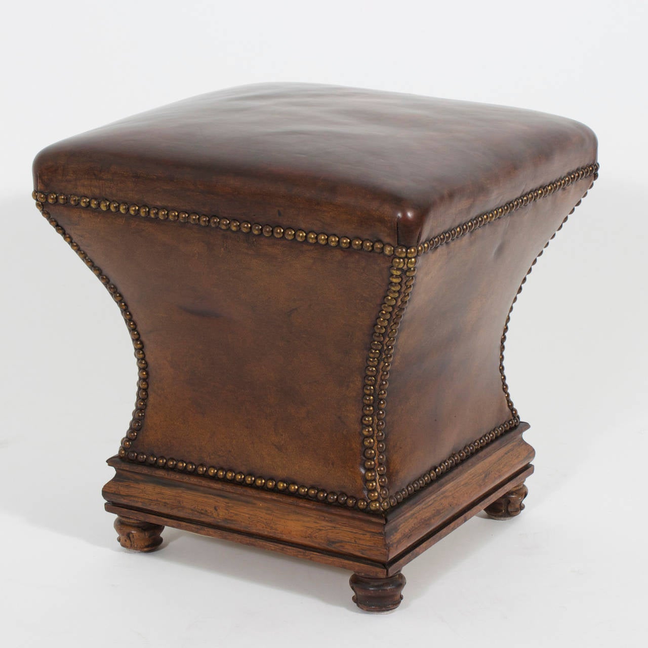 A pair of brown leather stools or ottomans clad in thick leather with brass tack concave outlines over a wooden base on turned and carved inverted tulip feet. Handsome, classic with a twist of gentlemen's club. Newly polished.