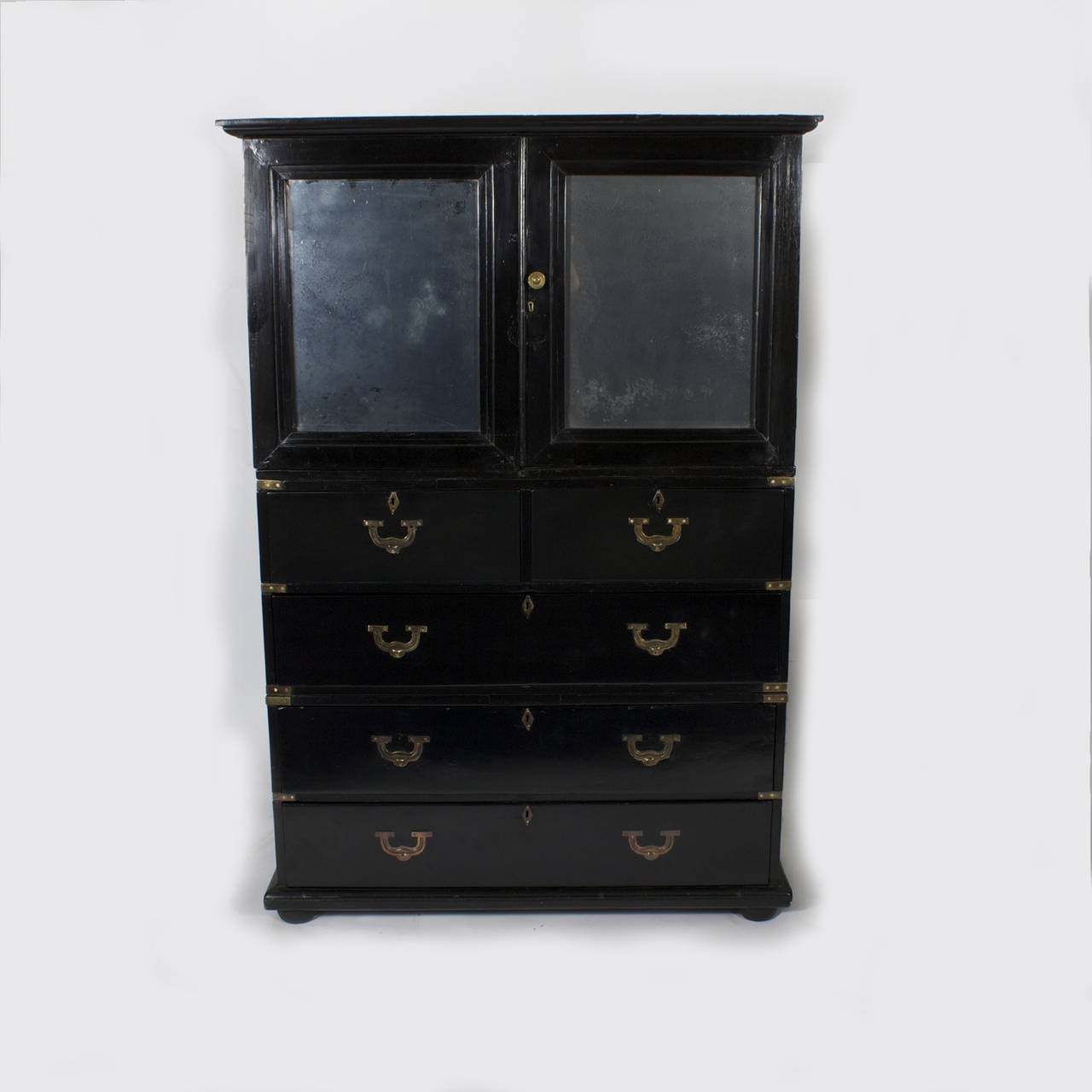 Unusual, ebonized combination cupboard or cabinet and chest of drawers, executed in Classic Campaign form. Two paneled mirror doors with time worn distress over four drawers with recesses brass pulls, original brass side handles and corner brackets,
