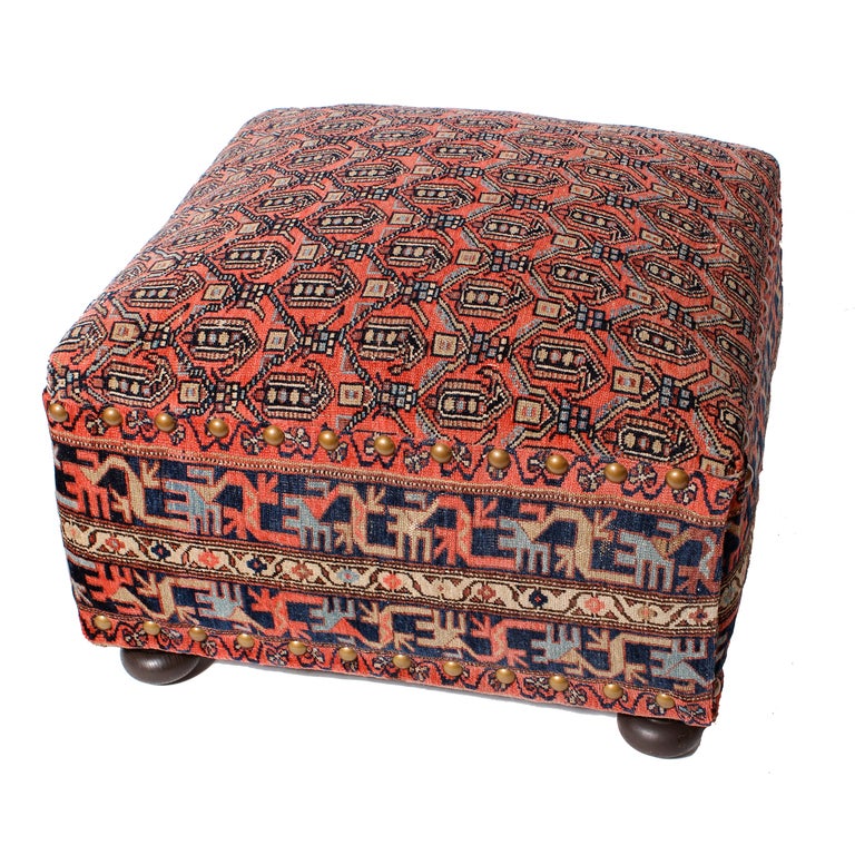A good example of a Shiraz oriental carpet fragment used as upholstery on a turned foot ottoman or footstool with brass tacking.
The field of the rug, is used on the ottoman top, while the carpet borders serve as the upholstered sides, with brass