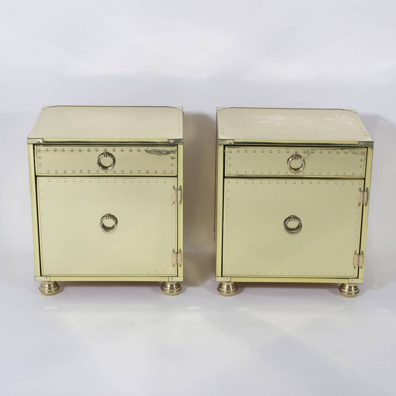 Pair of Mid-Century brass-plated nightstands or end tables, one drawer and door, with ring pulls, designed in a Campaign style and entirely clad in metal. Featuring faux rivet trim, turned brass feet and lots of storage, these stands coin a new