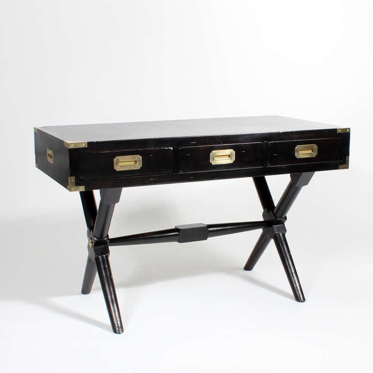 This is a very robust black stained pine 3 drawer campaign style desk with turned x base legs, and middle stretcher. Brass strapping, recessed hardware and decorative brass ring decorate the case. Campaign carrying handles and a 3 paneled back, make