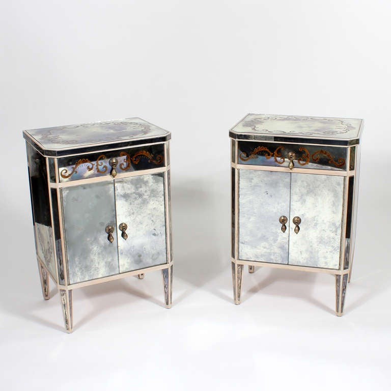 A pair of mirrored night stands, commodes or tables, with curlicue decorated tops, drawers and ends, banded by ivory painted wood, single drawers over double doors, with drop pulls. The mirrors have aged fantastically with irregular clouding