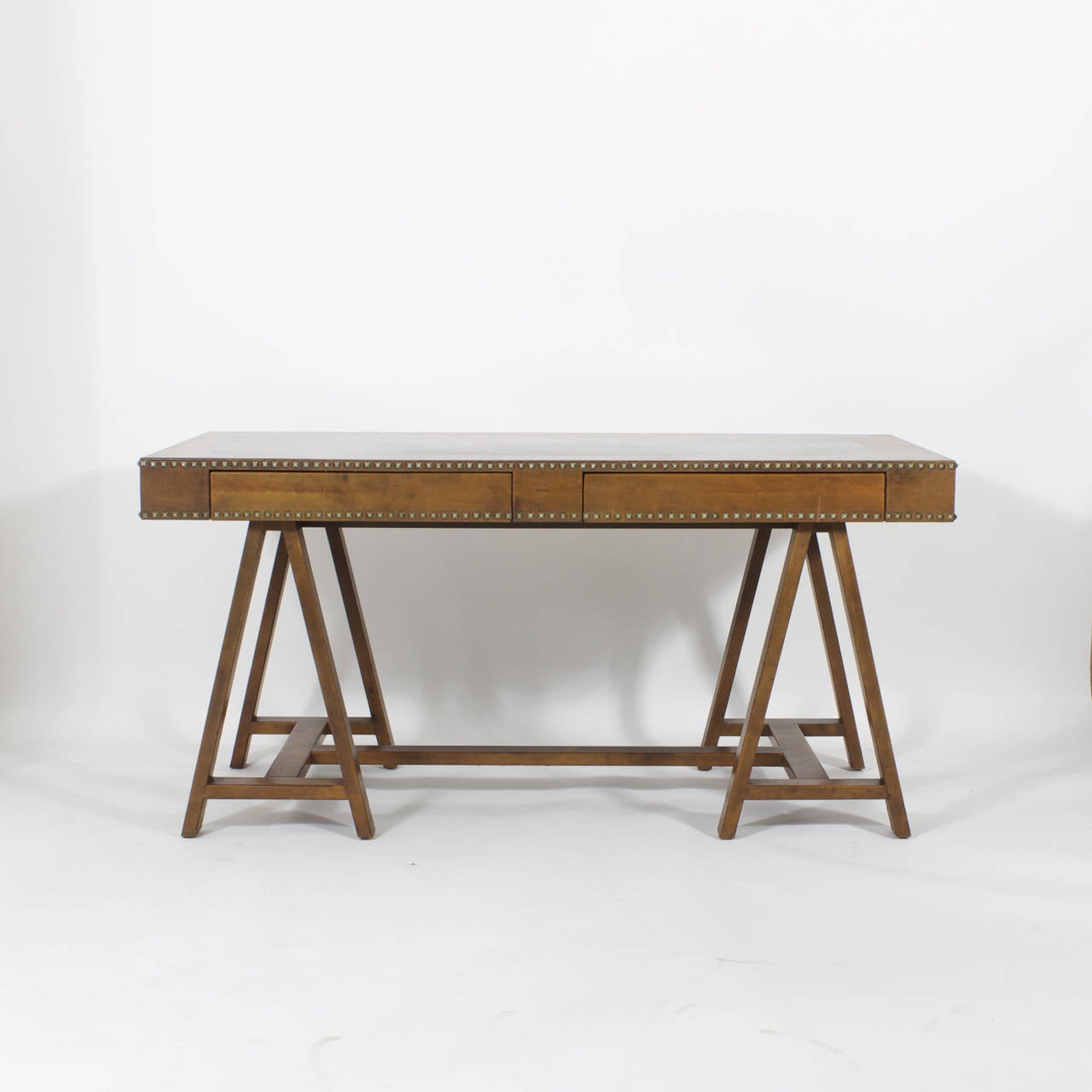 This two drawer, campaign style writing desk or table has classic lines with sawhorse legs. The desk, from top to legs is entirely clad in warm brown leather and trimmed with very decorative square brass tacks. Traditional meets modern in a grand