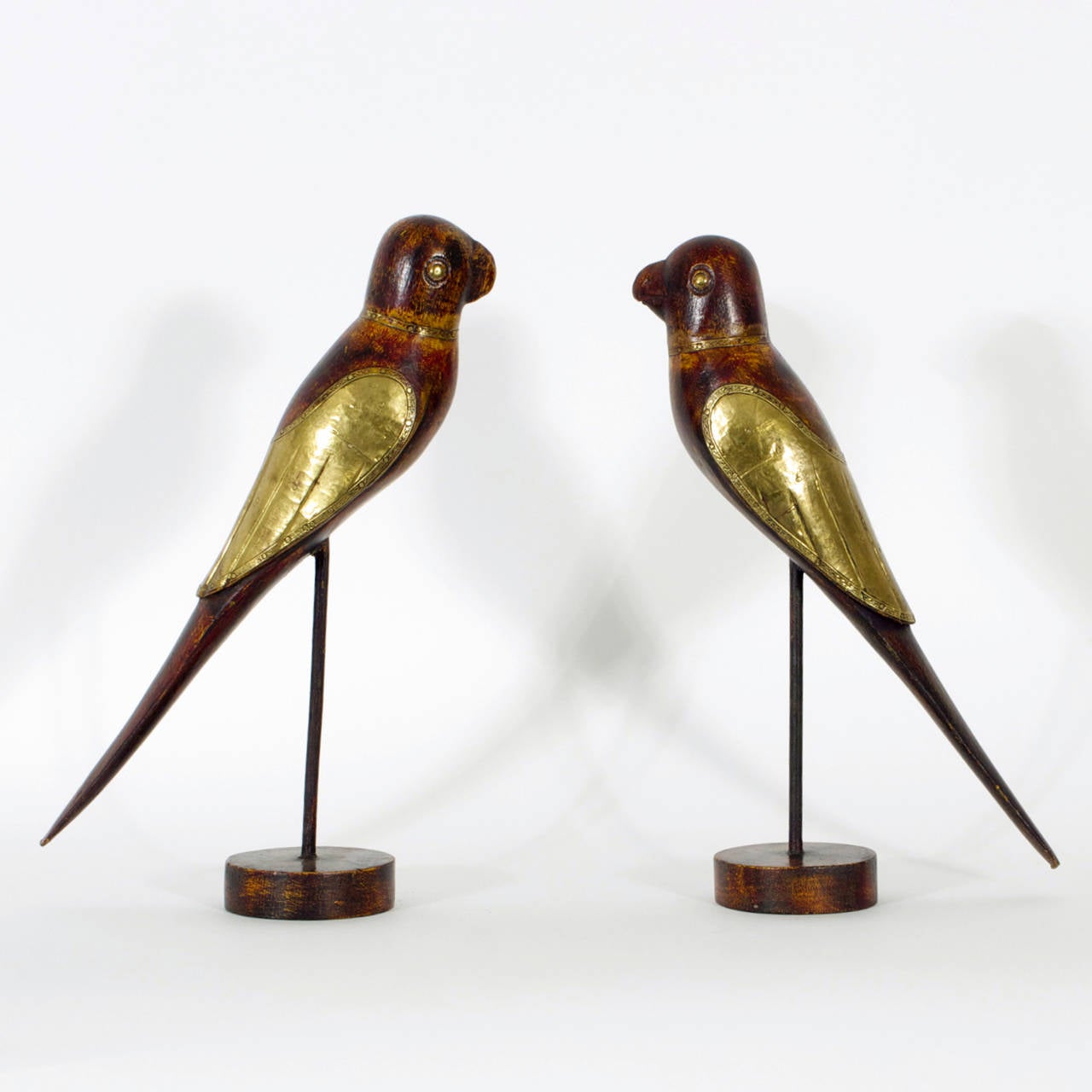Amusing pair of carved wood birds or parrots each having hammered and decorated brass wings, collars and eyes, all in warm worn paint. Mounted on metal stems supported on round wood bases. Rustic and soulful. Due to the hand carved nature, the birds