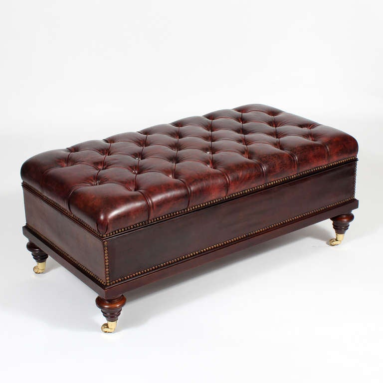 A leather covered ottoman with turned wood legs terminating in brass casters, with a tufted leather top, the whole outlined with brass tacks. The lid lifts for storage. A wonderful old and decorative piece that evokes Ralph Lauren, and everything