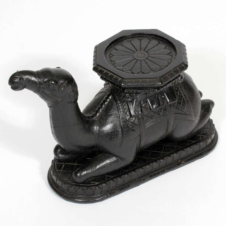 A rare size and form Anglo Indian carved and ebonized camel pedestal or vase holder. A camel in the lying position, with legs tucked under, dressed with an incised carved blanket and saddle, topped with an octagonal shaped vase holder. Adds an