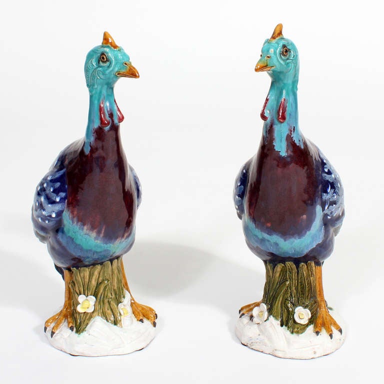 A pair of large glazed terracotta guinea fowls, with bold and bright colors, in blues and purples, standing on floral decorated trunks and white bases. Signed with a geometric mark on the bases, probably from Spain. Large in size and dynamic in