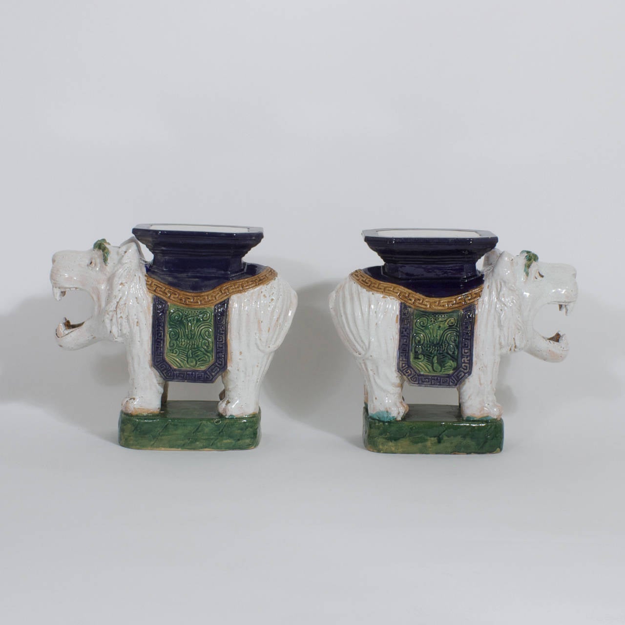 Whimsical pair of roaring lion tables or garden seats made of terra cotta then painted and glazed in white, green, blue and brown. These comical cats have been trained to work and wear saddles with pedestals. Perfect use for drink stands, plant