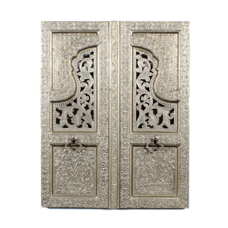 A pair of Indian silver plated panels, carved to look like shutter doors. Foliate carvings cover the body of the shutters, with vases and flowers complimenting the lower panels. Complete with silvered ring pulls. A very decorative element to