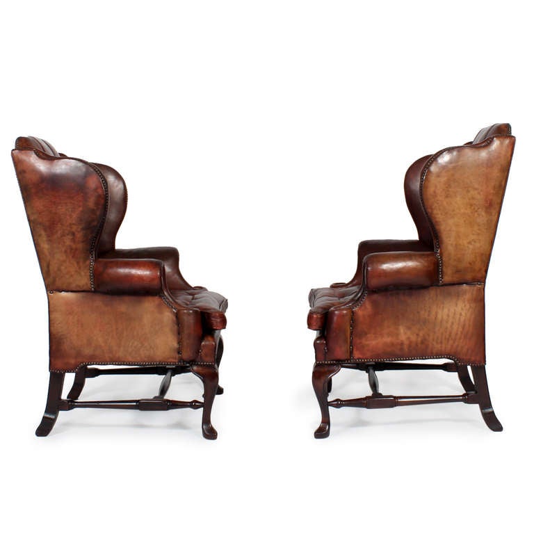 A pair of incredible well aged high back leather early 20th century wing or club chairs, with an exceptional Silhouette, rolled arms, cabriole legs, spade foot and H-turned stretchers all in varnished mahogany. Brass tacks accentuate the phenomenal