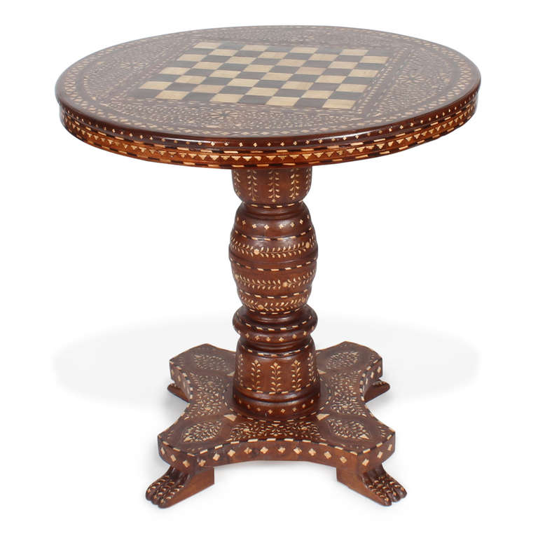 A large round Syrian games table with inlaid bone details, including the checkerboard top and extensive decorative foliate inlay throughout the table. A very bold turned pedestal base with a platform base and modified animal paw feet. Adds character