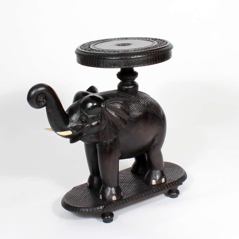 This is a very charming Anglo Indian stand or table, with a floral carved top and a full body carved elephant, trunk up, standing on a cross hatched base terminating with turned feet. Painted tusks and toe nails. We have a very similar smaller