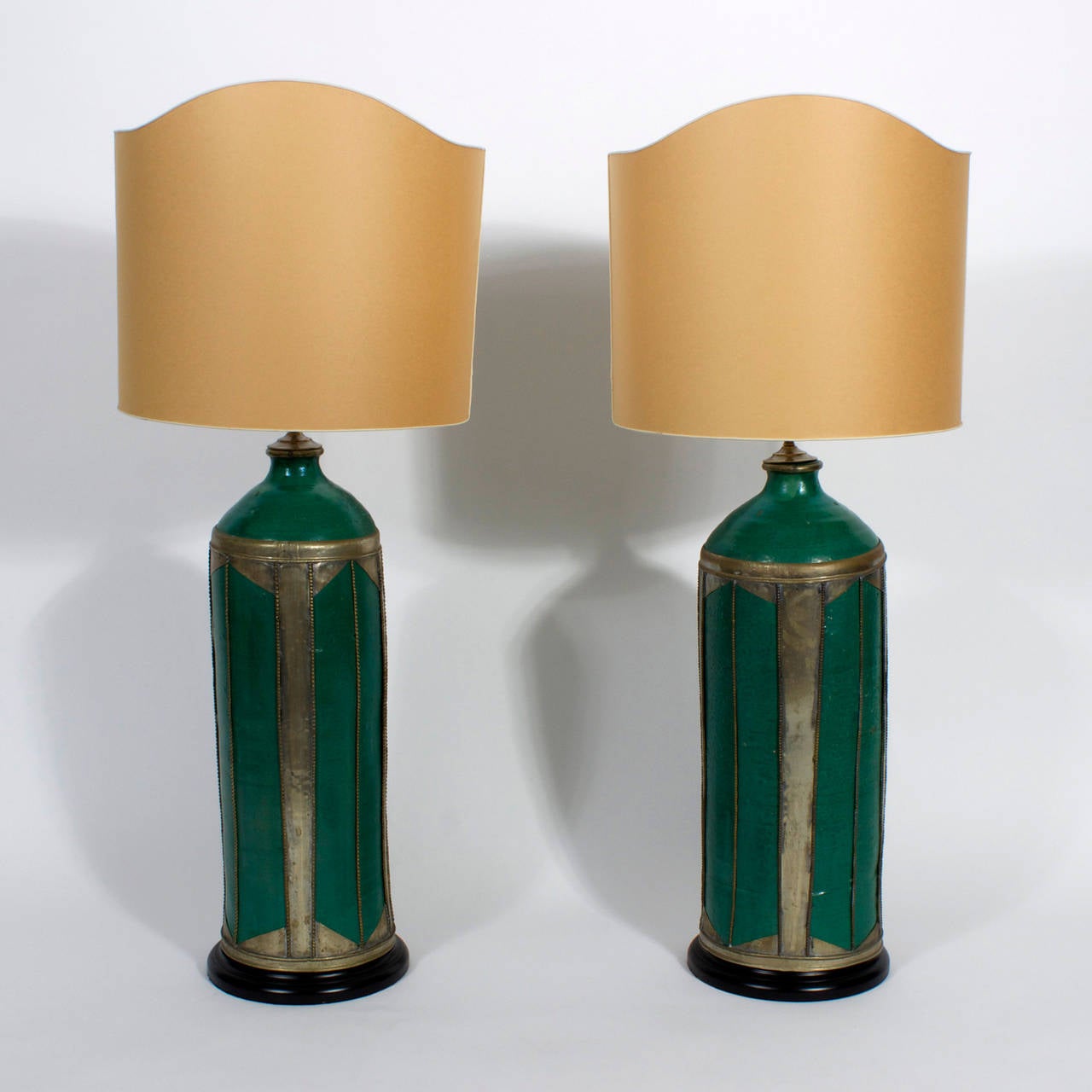 An interesting pair of custom green glazed pottery table lamps with applied hand hammered metal decoration and ebonized wood bases. These large scale and unique lamps have unusual custom shades, with scalloped tops and open backs, which allow the