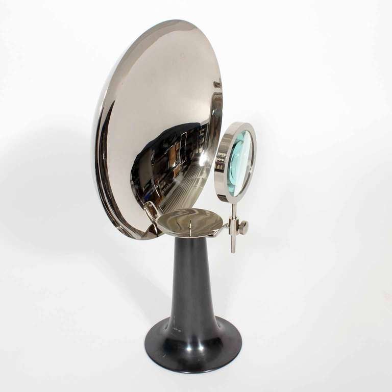 A very sculptural concave reflective candle holder , with a nickel plated pedestal base and disk back, and a plated reflective frontal surface, with a pricket candle holder, and adjustable magnifying glass. Not sure what these devices were used for