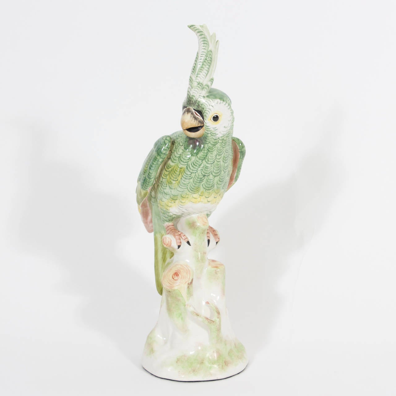 Italian porcelain parrot happily decorated with green, gold and white feathers, perched on a stout tree trunk.