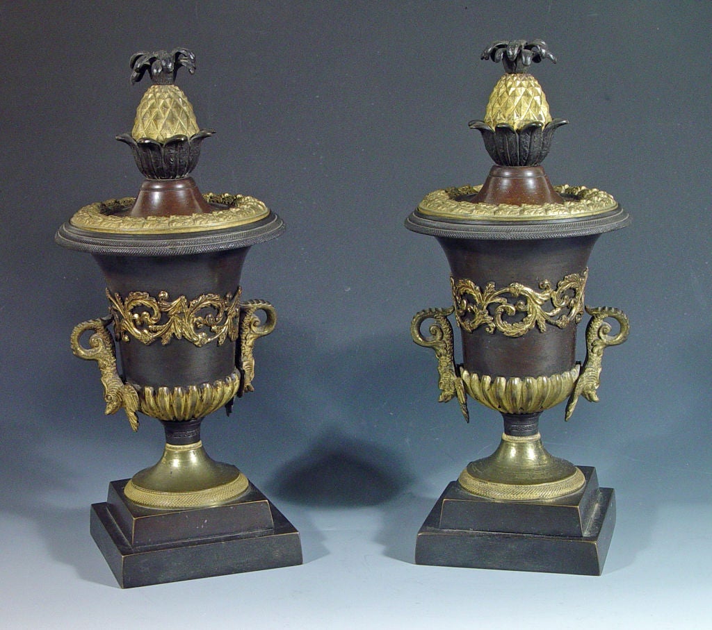 The bronze urns rest on an ormolu flaring foot which rests on a two step bronze square base. The ormolu handles in the form of dolphins are applied to each side and encircling each is a band of swirling ormolu leaves. The pierced top is surmounted