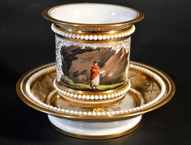 The cup, with Royal provenance, is finely painted with a figure of a woman with a large wicker basket looped over her left arm walking past large jagged rocks.  This scene is painted on a gilt vermilion ground.  The terminal of the gilt handle is in
