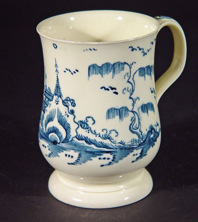 The early creamware tankard of bell-form is decorated with a Chinoiserie landscape of buildings along a river bank with trees and plants to either side with birds flying in the sky.  The loop handle with wonderfully crisp and fine applied terminals.