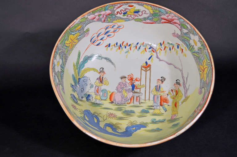 The bowl has a large panel on the interior well depicting Chinoiserie figures in a garden setting below a large shady tree with servants serving a sitting Mandarin figure who is sitting on a bench with a large tall wooden stand to his left adorned
