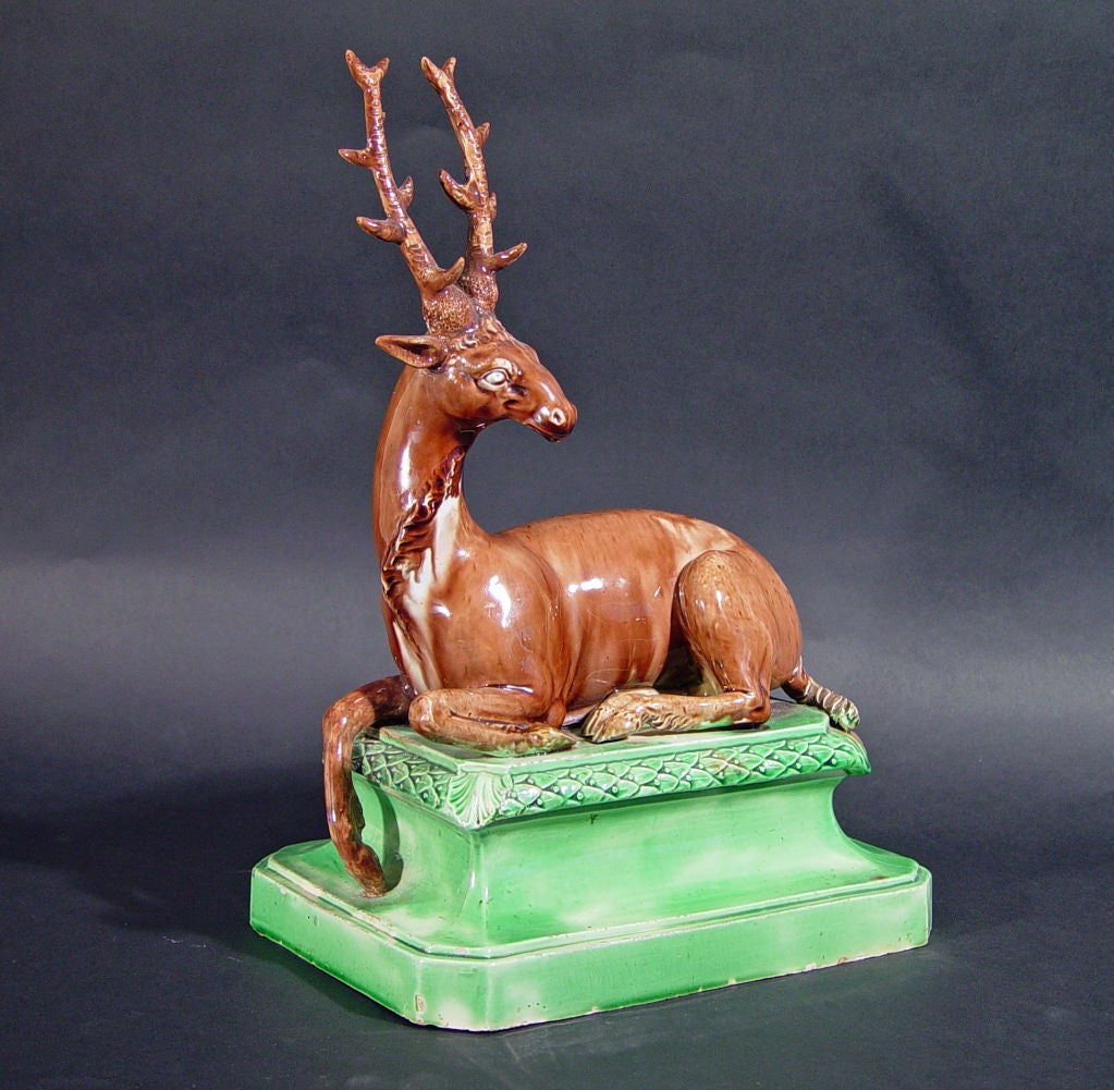 The stag with light brown coat revealing patches of white on her neck and eye, modelled recumbent with one foreleg draped over the side of the base, the green waisted base surrounded by a berried laurel leaf border and unglazed underneath.<br