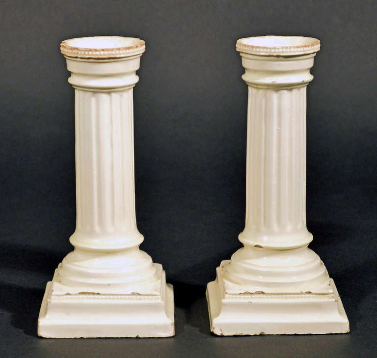 The pair of English 18th-Century creamware candlesticks are probably Leeds. These creamware candlesticks have a central reeded column rising from a stepped square base with beading around the top rim of the base and the edge of the candle