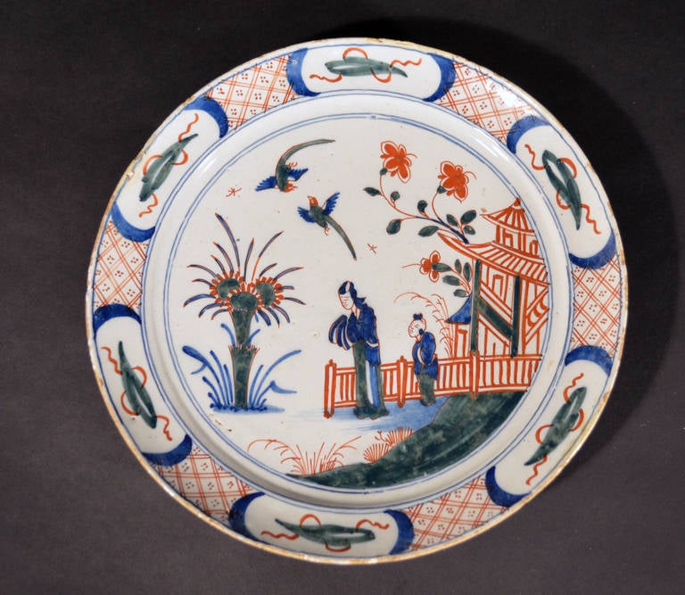 The pancake shaped plate is decorated with two Chinese figures in a garden landscape with red fence and pagoda in the background.

Mark: Grieksche A in red

Reference: Dutch Majolica & Delftware from the Edwin van Drecht Collection, Frits