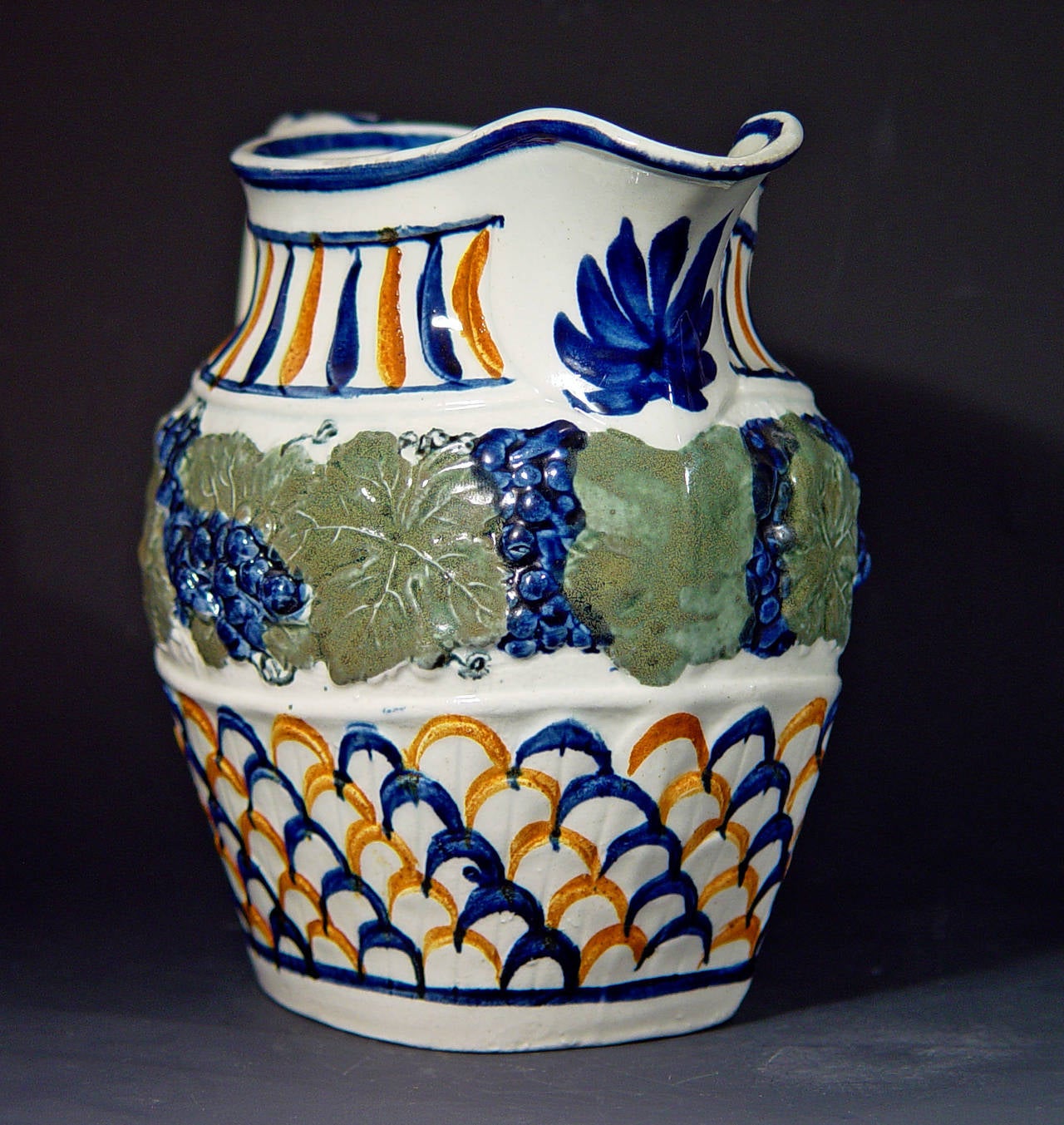 The jug is decorated in a pattern known as Vine & Scale with a moulded design of grapes and grape leaves surrounding the shoulder with a raised fish-scale design below in typical Pratt colors. The rim with an underglaze blue line and a large leaf in