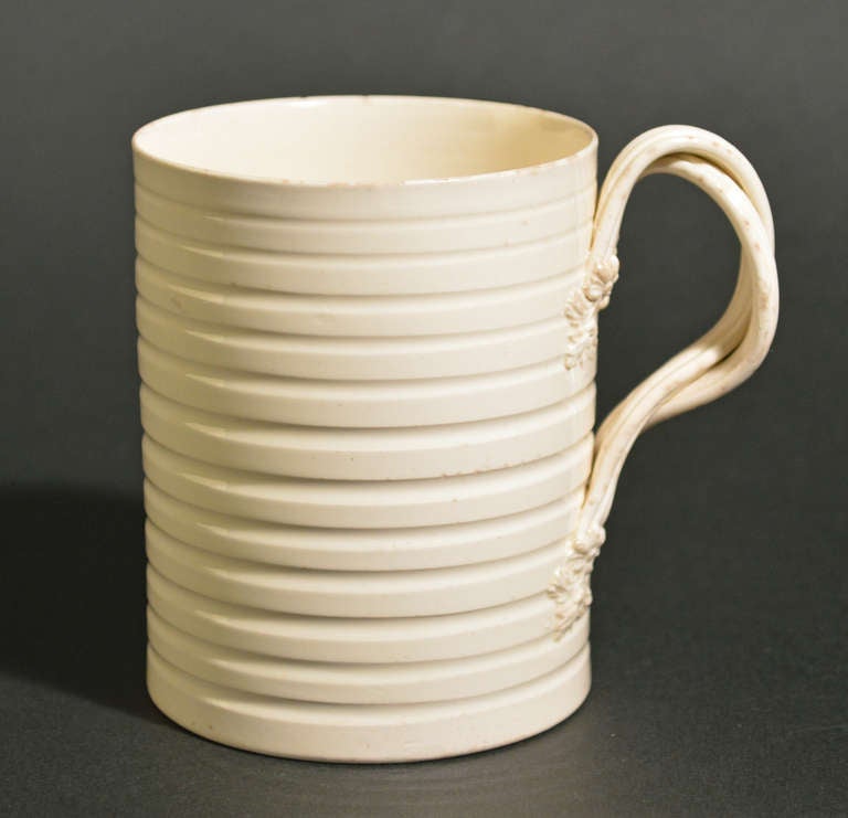 An English creamware pottery mug with a machine-turned body, featuring a twisted strap handle connected to the body of the mug with a flower and leaf terminal.