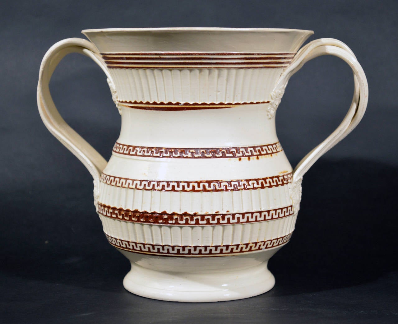 The body turned and rouletted with plain bands, reeding and a Greek key design with a brown slip ground. The large cup with two double twist reeded handles with oak leaf terminals.