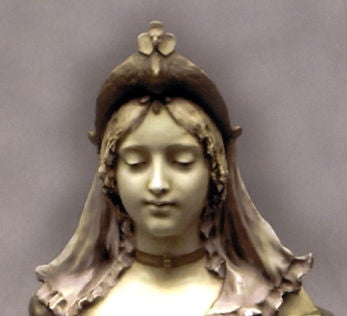 Young lady wearing a green gown and hair piece.  The bust sits on a pedestal base, all in one piece, which has a scene, in relief, of a female on horseback in a forest.