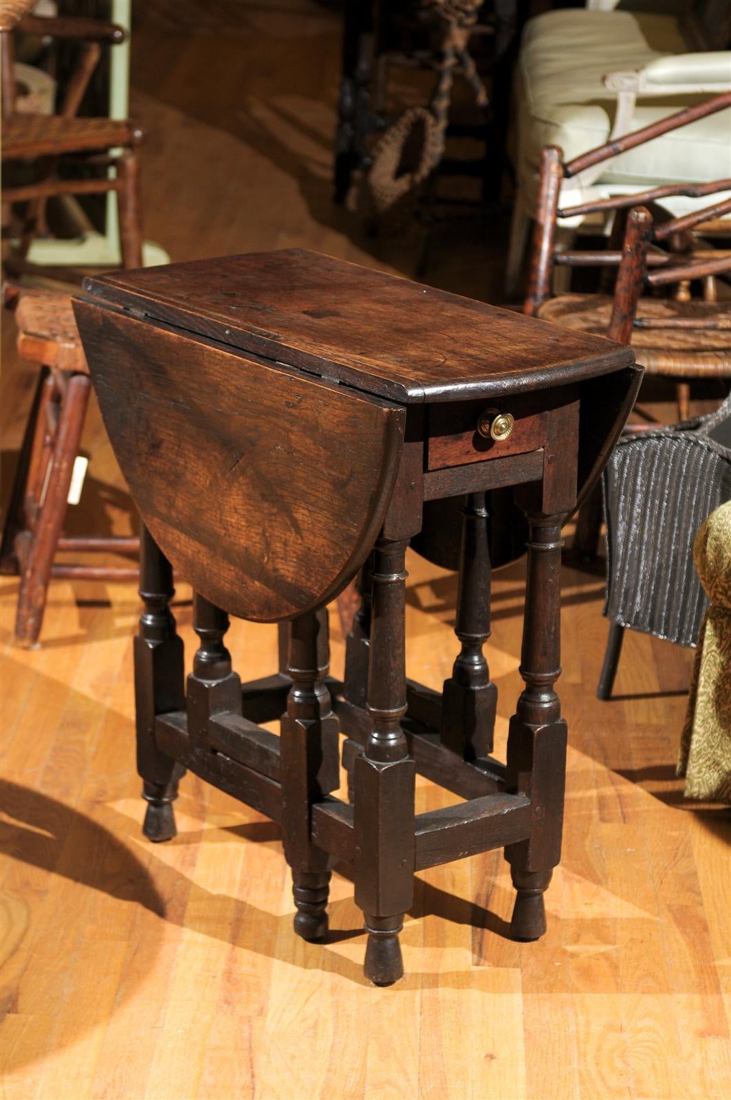 This is a wonderful somewhat primitive gate leg table with charm and patina. The edge of the table has a rounded profile. The table top is solid wood with a drawer in the center. The wood is dark English Oak with pegged construction.