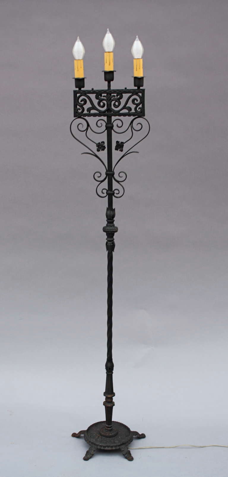 Classic wrought iron torchiere with three candle lights above decorative cast panel and pretty scrolls and flowers up stem.