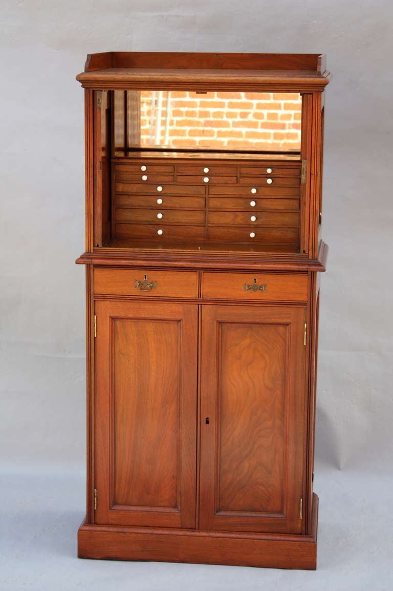 Tall & elegant dental or medical cabinet with highly carved doors which slide back into top portion of cabinet itself to reveal mirrored interior and fourteen tiny drawers. Bottom section features two standard drawers and large cupboard space with