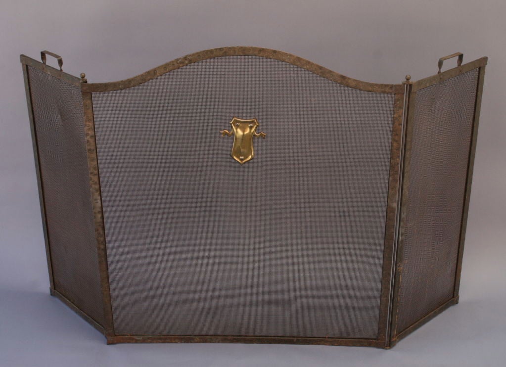 Gracefully arched hammered brass fire screen with crest or shield medallion at center; tri-fold design allows for size adjustment; handles for ease of movement