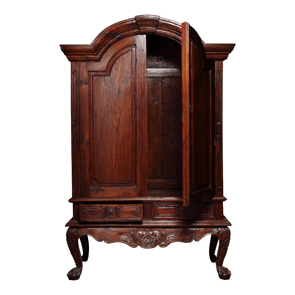 This large size Dutch colonial style Indonesian armoire from the 19th century features two doors over two drawers. The eye is immediately drawn to the lovely bonnet shaped arched pediment. The doors below are carved and show recessed panels that