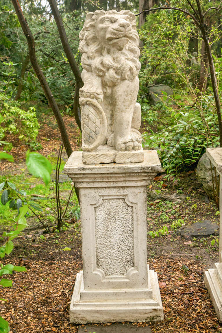 A pair of carved stone gate pier seated lions, looking in opposing directions, each fully-maned beast with one paw raised on a heraldic shield, Bavaria, early 19th century on an associated pair of composition stone plinths with inset panels, the