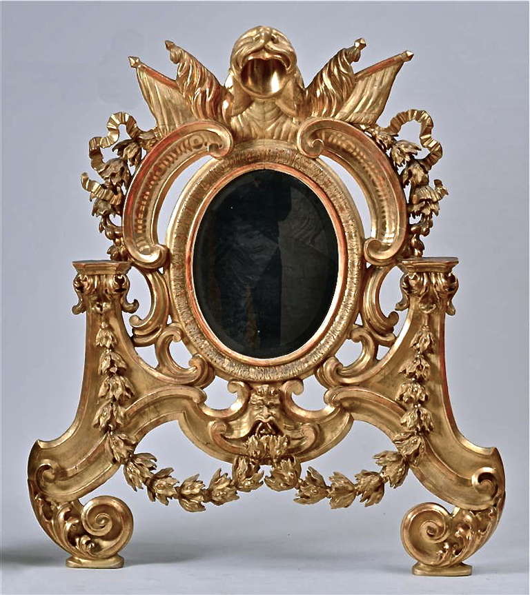 The oval beveled glass mirror plate surmounted flags and a lion pelt flanked by C-scrolls and ribbon swags, continuing to small brackets issuing berried laurel swags joined by a male mask, terminating in acanthus scrolls.