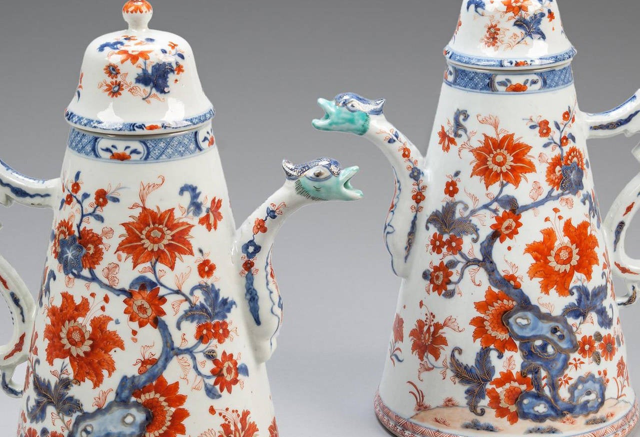 A pair of early 18th century Yongzheng conical porcelain chocolate pots and covers richly decorated in the Imari style, the cylindrical body with blue and white trailing flowers with rich orange peonies against a white ground in a stylized garden