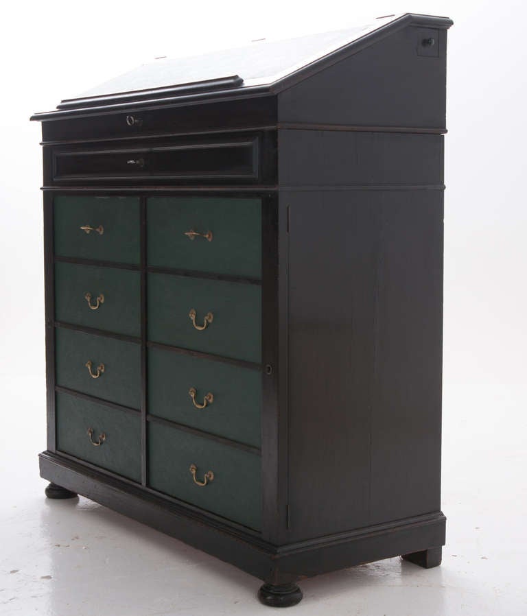 Continental notary's desk, featuring hinged slant top with green writing surface and paper prop. To right side is a small square drawer which pulls out to be a pencil holder. Full length drawer sits above the faux cabinet doors which open for