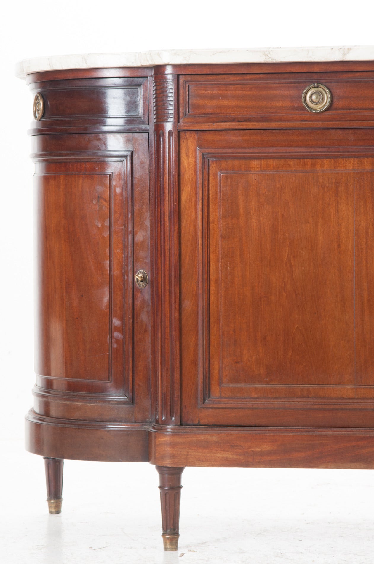 A classic Louis XVI style mahogany demilune buffet / enfilade with its original white marble top. Four spacious drawers and four doors allow for  lots of storage. The pronounced and fluted corners are perfect on  this detailed yet simple buffet. It