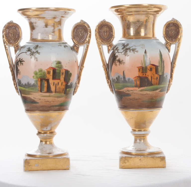This pair of antique French, Empire style, polychrome porcelain urns are exquisite and in superb condition. Rich colors of red, green, yellow and gold. On each handle is a carved male's face. The scenery is romantic, with a painted chateau on one