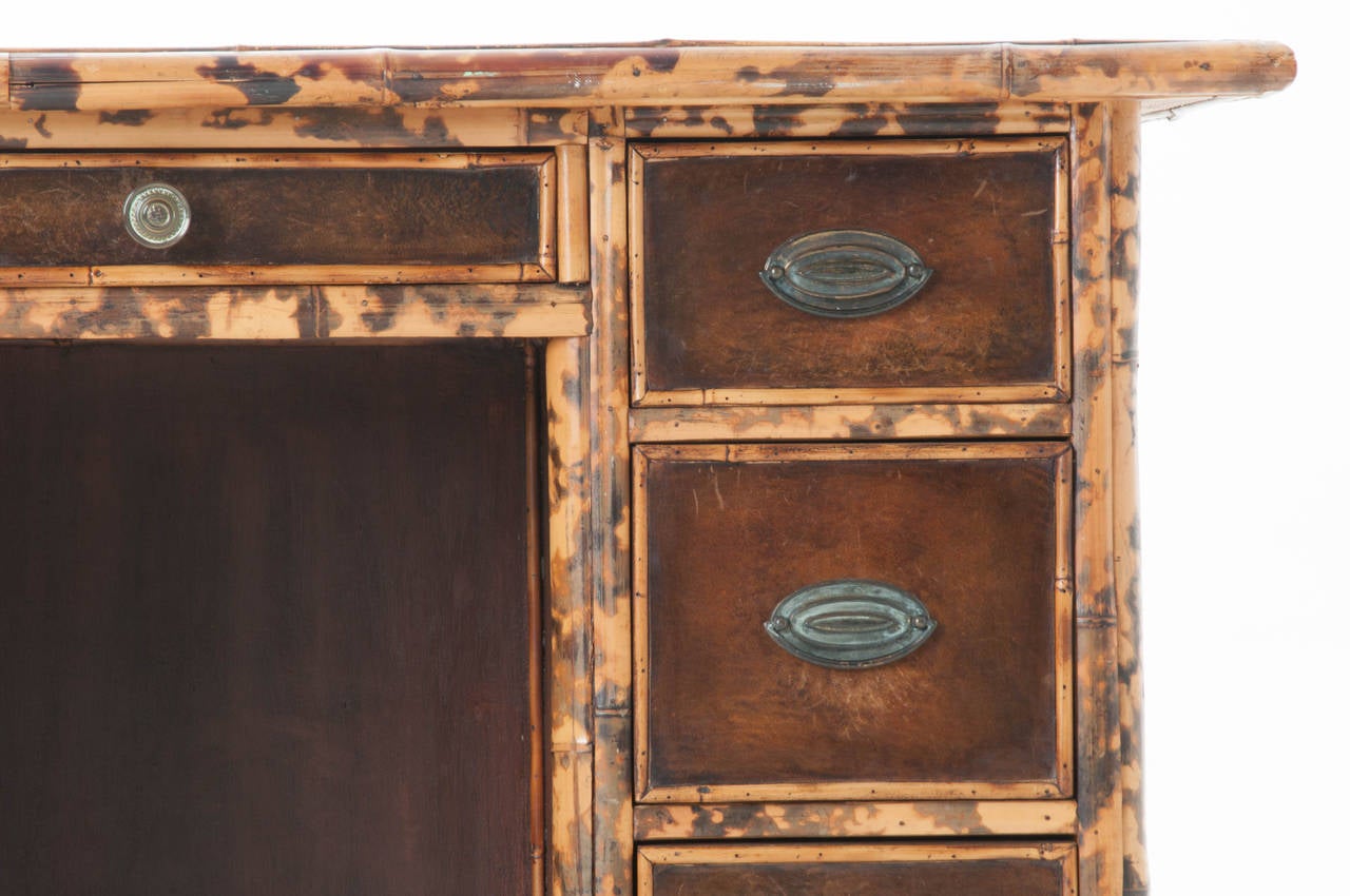 English bamboo and leather desk from the early 1900's. Embossed Greek key edging on leather top, front and sides, the 5 drawers have simple leather fronts has replaced the original sea grass. 
Knee height is 24.13