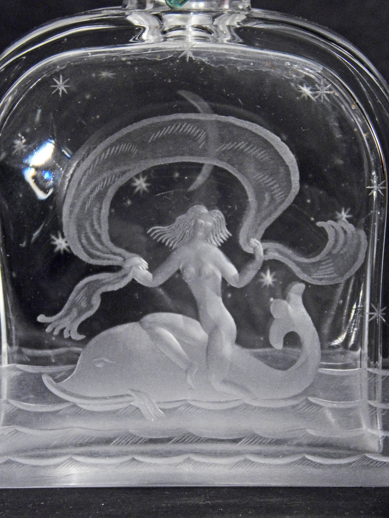 A tour de force of glass engraving, by the famed Orrefors glass company in Sweden, this perfume bottle depicts a nude mermaid riding a dolphin, holding a billowing length of drapery overhead. The Orrefors designer and engravers missed no detail,
