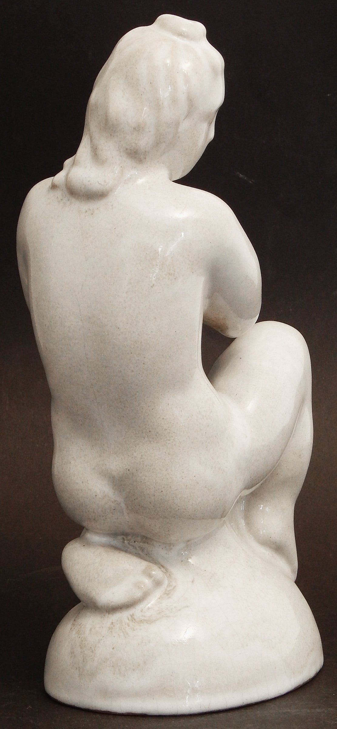 Finished in a soft ivory glaze with subtle shifts to dove grey, this lovely, placid nude is depicted in a seated position with her hands clasped and her gaze looking into the distance, the subject deep in thought. Unsigned, though marked with a 